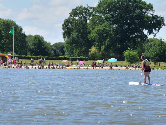 Une femme fait du stand up paddle sur le plan d'eau de La Rincerie. Dans le fond on voit des baigneurs sur la plage.