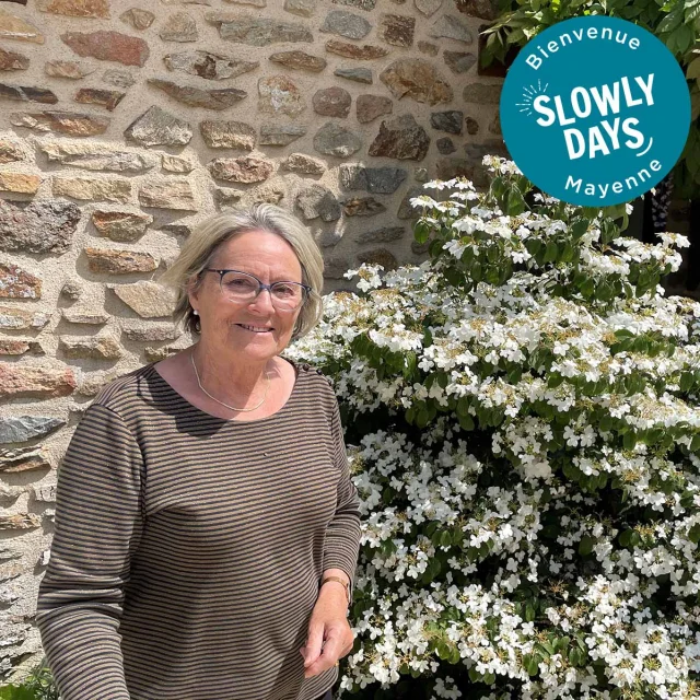 Photo de Françoise Beuvier devant un mur en pierre de sa maison. A l'arrière plan on voit également un arbuste en pleine floraison. Dans le coin supérieur droit on voit l'estampille ronde bleue sur laquelle est écrit en blanc au centre Slowlydays. En haut on lit Bienvenue et en bas Mayenne