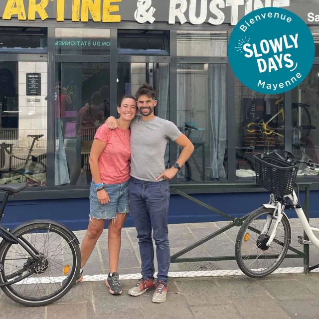 Marine et Damien posent souriants devant leur établissement Tartine & Rustine à Laval. Ils sont , entourés de vélos. Un badge Bienvenue SlowlyDays Mayenne apparaît en haut à droite de l'image.